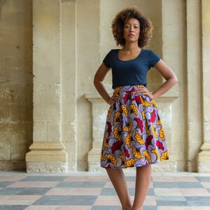 May include: A woman with curly hair is wearing a navy blue top and a colorful skirt with a floral pattern. The skirt has yellow, red, and purple flowers on a white background. She is standing in front of a stone wall.
