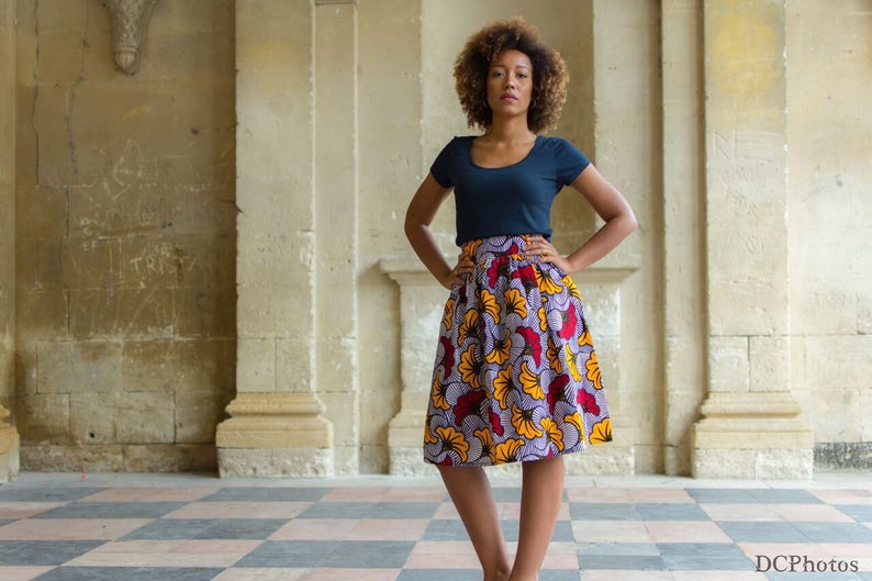May include: A woman with curly hair is wearing a navy blue top and a colorful skirt with a floral pattern. The skirt has yellow, red, and purple flowers on a white background. She is standing in front of a stone wall.