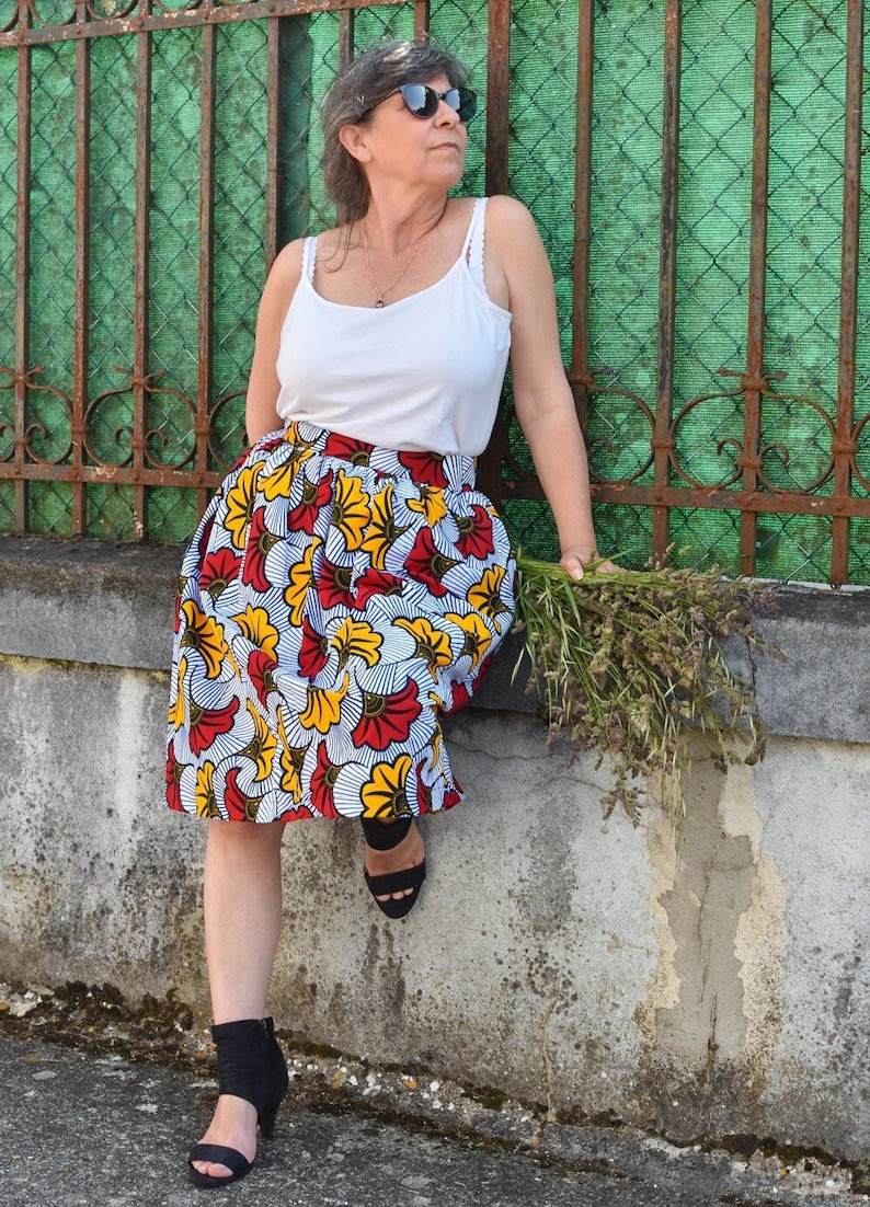 May include: A woman wearing a white tank top and a colorful floral skirt with red, yellow, and white flowers. She is wearing black sandals and sunglasses. She is holding a bouquet of dried flowers.