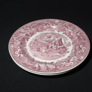 May include: A pink and white ceramic plate with a floral border and a central image of a ship sailing into a harbour.