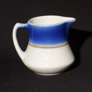 Op de afbeelding: Een klein wit keramiek melkkannetje met een blauwe en gouden band rond de bovenkant. Het melkkannetje heeft een handvat en een tuit.