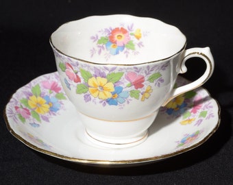 May include: A white teacup and saucer with a floral pattern in shades of pink, yellow, blue, and purple. The cup and saucer have a gold trim.