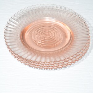 Puede incluir: Un conjunto de platos de vidrio rosa apilados. Cada plato presenta un borde acanalado y un centro circular y estriado. Los platos son de un tono rosa suave y est&aacute;n apilados. Se presentan sobre una superficie blanca.