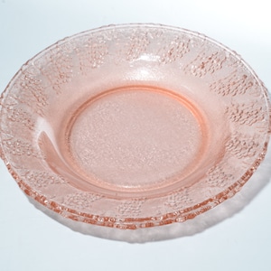 Könnte beinhalten: Eine rosa Glasschale mit strukturierter Oberfläche und einem dekorativen Traubenmuster am Rand. Die Schale ist rund und hat eine geringe Tiefe, geeignet zum Servieren von Salaten oder Desserts. Die Schale ist aus Glas.