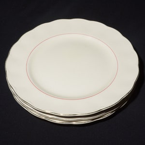 May include: A stack of four white ceramic dinner plates with a red rim and a gold trim. The plates have a scalloped edge.