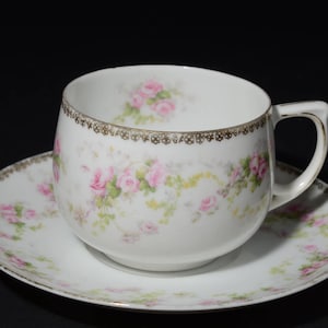 Puede incluir: Una taza de té y un platillo blancos con un patrón floral de rosas rosadas y ribete dorado.