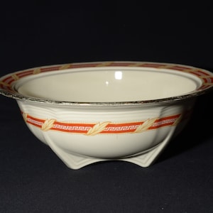 May include: A white ceramic bowl with a red and gold geometric design. The bowl has a wide rim and a flared base with three feet.
