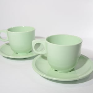 Könnte beinhalten: Zwei hellgrüne Keramik-Teetassen und Untertassen. Die Tassen haben eine abgerundete Form und einen kleinen Henkel. Die Untertassen sind etwas größer als die Tassen und haben einen flachen Boden.