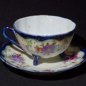 Vintage doorschijnend theekopje en schoteltje op voetjes, gemaakt van fijn porselein met een bloemenboeket van rozen en een handbeschilderde kobaltblauwe rand.