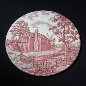 May include: A white ceramic plate featuring a red and white design of a cottage and windmill in a countryside scene.