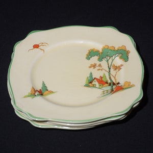 Può includere: Una pila di piatti quadrati color crema con un bordo verde smerlato. Ogni piatto presenta un paesaggio con una casa, alberi e un sole. I piatti sono impilati su una superficie scura.