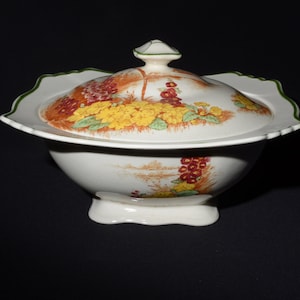 May include: A white ceramic serving bowl with a lid. The lid and bowl are decorated with a floral pattern in yellow, orange, and red. The rim of the lid has a green trim. The bowl sits on a white base.