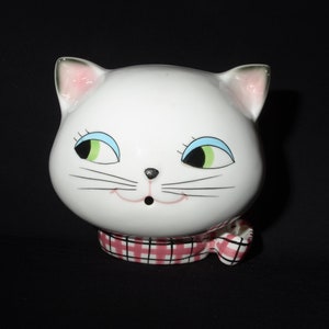 May include: White ceramic cat figurine with green eyes and a pink and white checked scarf.
