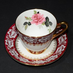 May include: A white teacup and saucer with a red and gold floral design. The teacup has a single pink rose with green leaves painted on the inside. The saucer has a gold rim and a white floral design on a red background.
