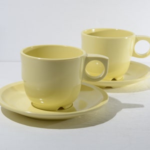 Può includere: Due tazze da caffè in ceramica gialla con piattini abbinati. Le tazze hanno una forma arrotondata e un piccolo manico.