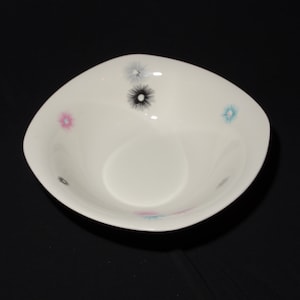 Könnte beinhalten: Eine weiße Keramikschüssel mit quadratischer Form und leicht abgerundetem Rand. Die Schüssel hat ein schlichtes Design mit fünf kleinen, stilisierten Blumen in Rosa-, Blau- und Schwarztönen.