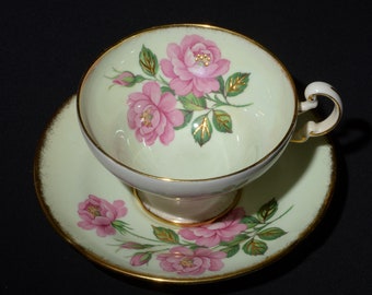 May include: A vintage teacup and saucer set with a delicate floral design. The cup and saucer are both pale green with a gold rim and feature pink roses with green leaves.