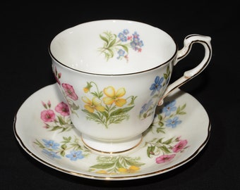 May include: White teacup and saucer set with a floral pattern featuring yellow, pink, and blue flowers. The teacup has a gold rim and handle, and the saucer mirrors the floral design. The set is likely made of porcelain.