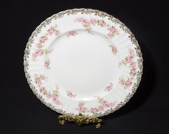 May include: A white porcelain dinner plate with a gold rim and a delicate pink rose pattern. The plate is on a gold metal stand.
