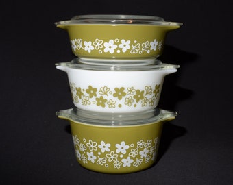 Puede incluir: Una pila de tres fuentes redondas de vidrio para horno con tapa. Las fuentes superior e inferior son de color verde oliva con un patrón floral blanco, mientras que la central es blanca con un patrón floral verde oliva. Tienen dos asas.
