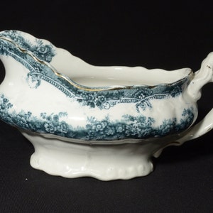 May include: A white porcelain gravy boat with a green and gold floral design. The boat has a handle and a scalloped edge.