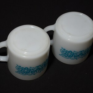 May include: Two white ceramic mugs with a turquoise floral design. Each mug has a rounded shape, a handle, and a floral pattern around the base. The mugs are set against a black background.