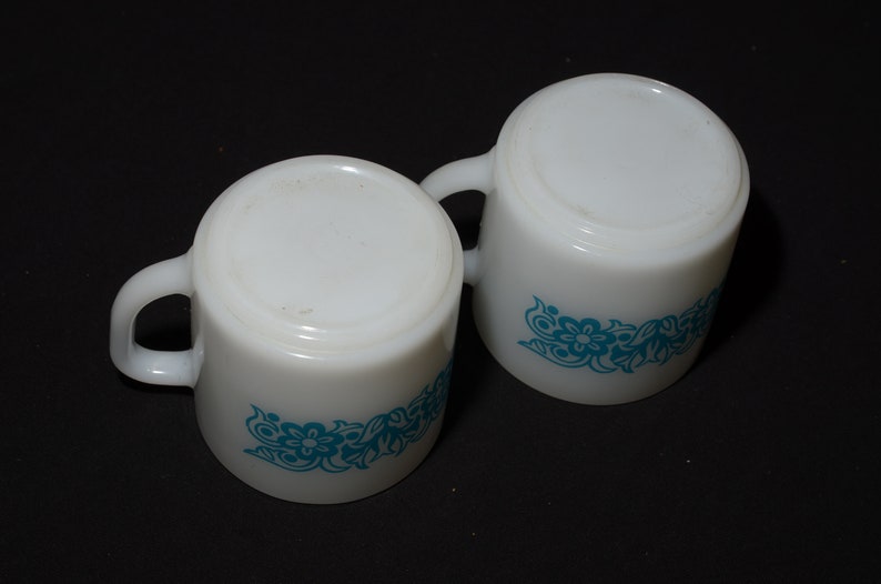 May include: Two white ceramic mugs with a turquoise floral design. Each mug has a rounded shape, a handle, and a floral pattern around the base. The mugs are set against a black background.