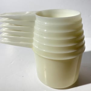 Puede incluir: Un juego de tazas medidoras apilables de color blanquecino. El juego incluye una taza grande con un exterior acanalado y una serie de cucharas medidoras rectangulares más pequeñas unidas al lateral. Las tazas y cucharas son de plástico translúcido.