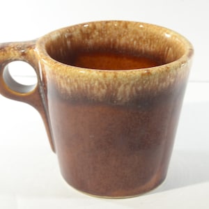 May include: A brown ceramic mug with a unique handle design. The mug has a dark brown base that fades to a lighter brown and tan rim. The handle features a circular opening for easy gripping. The mug is likely for coffee or tea.