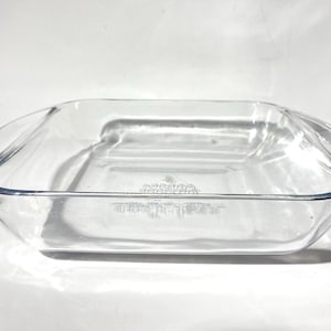 May include: A clear, rectangular glass baking dish with rounded corners. The dish is empty and sits on a white surface, with shadows cast across it. The dish appears to be made of durable glass.