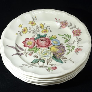 May include: A collection of white ceramic plates with a scalloped rim. Each plate is decorated with a floral pattern in shades of pink, yellow, and purple. The plates are stacked on a black surface.