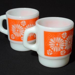 Puede incluir: Dos tazas de cerámica blancas con diseños florales naranjas. Las tazas tienen forma cuadrada con asa y base apilada. El diseño floral presenta grandes margaritas blancas y formas de flores más pequeñas. Las tazas están sobre un fondo negro.