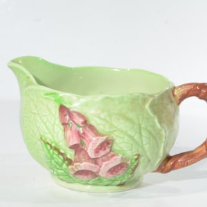 Puede incluir: Una jarra de cerámica verde con un asa marrón en forma de rama. La jarra está decorada con un diseño floral de digitales rosas y hojas verdes.