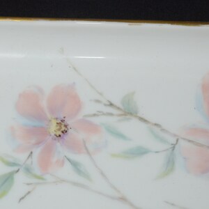 May include: A white rectangular dish with a gold rim, adorned with hand-painted pink flowers, green leaves, and delicate gray branches. The floral design adds a touch of elegance to this decorative piece.