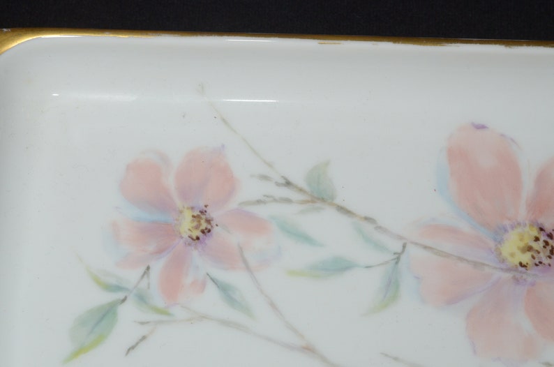 May include: A white rectangular dish with a gold rim, adorned with hand-painted pink flowers, green leaves, and delicate gray branches. The floral design adds a touch of elegance to this decorative piece.