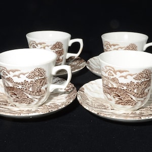 Puede incluir: Cuatro tazas de té blancas con platillos a juego. Cada taza y platillo presenta un diseño de paisaje marrón con un puente y árboles. Las tazas tienen asas curvas y se asientan sobre platillos redondos.