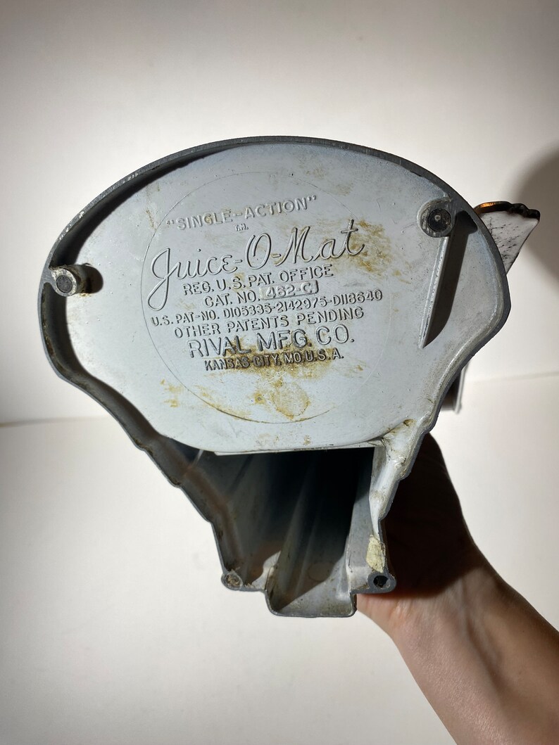 May include: A vintage silver-colored "Juice-O-Mat" single-action juicer with the text "REG. U.S. PAT. OFFICE CAT. NO. 462-CJ U.S. PAT-NO. DI05335-2142975-D118840 OTHER PATENTS PENDING RIVAL MEG. CO. KANSAS CITY, MO. U.S.A."