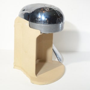 May include: A chrome and beige manual citrus juicer with a lever on the side. The juicer is made of metal and has a beige plastic base.