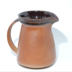 May include: A ceramic jug with a rounded body and a curved handle. The jug is a warm, earthy brown colour with a darker brown rim and interior. It has a spout for easy pouring and is likely used for serving beverages.