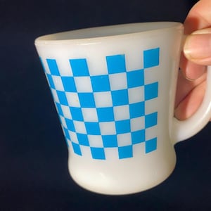 May include: White ceramic mug with a blue and white chequered pattern. The mug has a curved shape and a handle. The mug is held by a hand. The mug is a retro style.