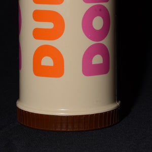May include: A beige Dunkin' Donuts travel mug with orange and pink lettering. The bottom of the mug is brown with a ridged texture. The mug is cylindrical and appears to be made of plastic.