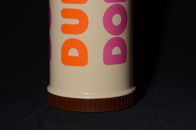 May include: A beige Dunkin' Donuts travel mug with orange and pink lettering. The bottom of the mug is brown with a ridged texture. The mug is cylindrical and appears to be made of plastic.