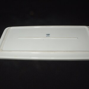 May include: A rectangular white ceramic serving platter, viewed from the underside. The platter has a slightly raised edge and a recessed area in the center. A small blue emblem is visible near the center.