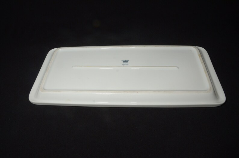 May include: A rectangular white ceramic serving platter, viewed from the underside. The platter has a slightly raised edge and a recessed area in the center. A small blue emblem is visible near the center.