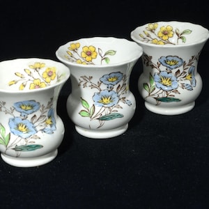 Op de afbeelding: Drie witte keramische vazen met een uitlopende bovenkant en een bolvormig lichaam. Elke vaas is versierd met blauwe en gele bloemmotieven en groene bladeren. De vazen zijn gerangschikt op een zwart oppervlak.