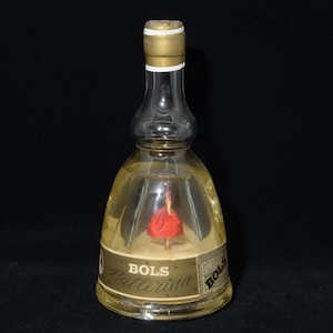May include: A vintage Bols Ballerina bottle with a clear glass body and a gold-colored cap. Inside, a small ballerina figure in a red dress is visible. The bottle has a label with the text "Bols Ballerina".