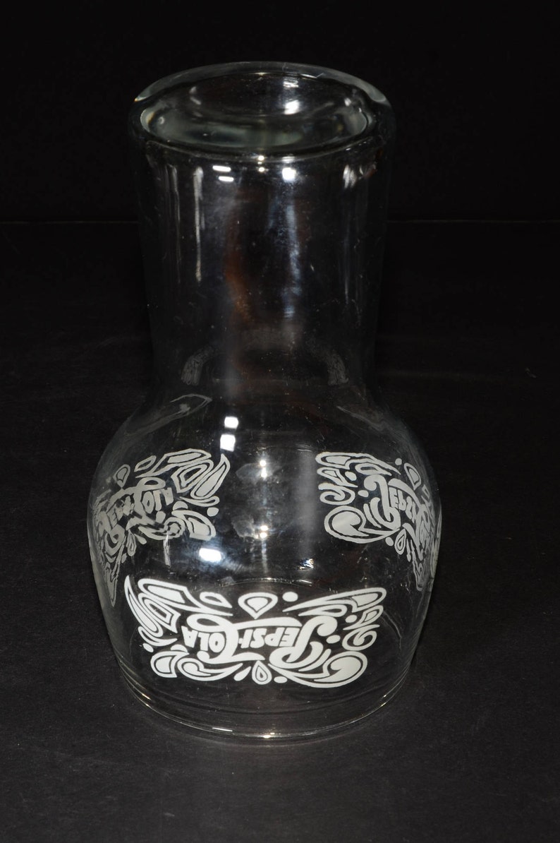 May include: A clear glass pitcher with a white etched design of the Pepsi-Cola logo and script. The design features stylized swirls and the words "Pepsi-Cola".