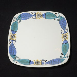 Può includere: Un piatto quadrato in ceramica bianca con un design di pesci blu e verdi attorno al bordo. I pesci hanno contorni neri e accenti gialli.