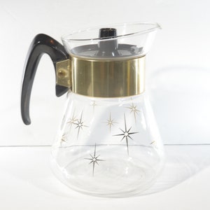 Może przedstawiać: Szklany dzbanek do kawy z czarnym plastikowym uchwytem i złotą metalową obręczą wokół górnej części. Dzbanek ma wzór gwiazdy po bokach.