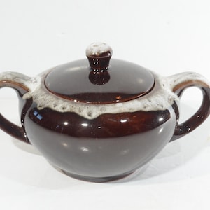 May include: A brown ceramic sugar bowl with a lid and two handles. The bowl has a glossy finish with a lighter-coloured trim around the rim and on the handles. The lid has a small knob.